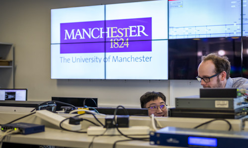 Two men looking at electronic equipment on a table, while a large screen behind contains The University of Manchester logo