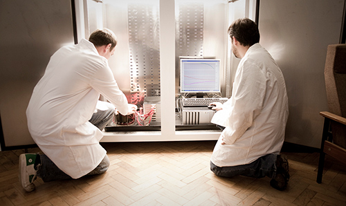 Two researchers in lab coats knelt on floor, adjusting PC wires